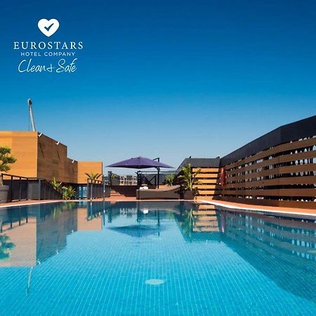Eurostars Palace Κόρδοβα Εξωτερικό φωτογραφία The photo shows an outdoor swimming pool with clear, blue water, reflecting the blue sky above. The pool area is surrounded by modern architectural features, including wooden slats and potted plants. There is also a purple umbrella providing shade. T