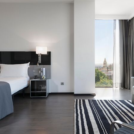 Eurostars Palace Κόρδοβα Εξωτερικό φωτογραφία The photo shows a modern hotel room featuring a large bed with white linens and dark cushions, paired with a sleek nightstand. There is a contemporary chair and a small table nearby, along with a striped area rug on the floor. A large window allows n