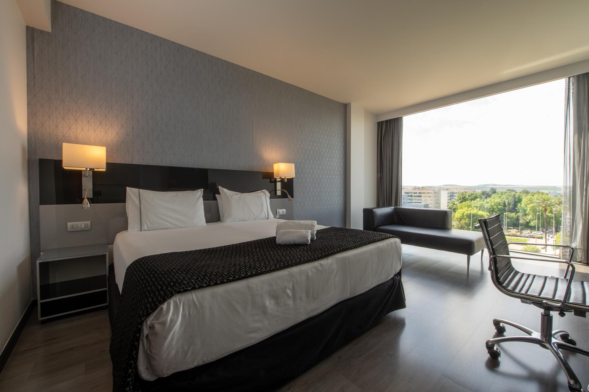 Eurostars Palace Κόρδοβα Εξωτερικό φωτογραφία The photo shows a modern hotel room featuring a large bed with white linens and a dark patterned throw. On either side of the bed, there are stylish wall-mounted lamps. A contemporary desk chair is positioned at a small desk, and there’s a sofa near 
