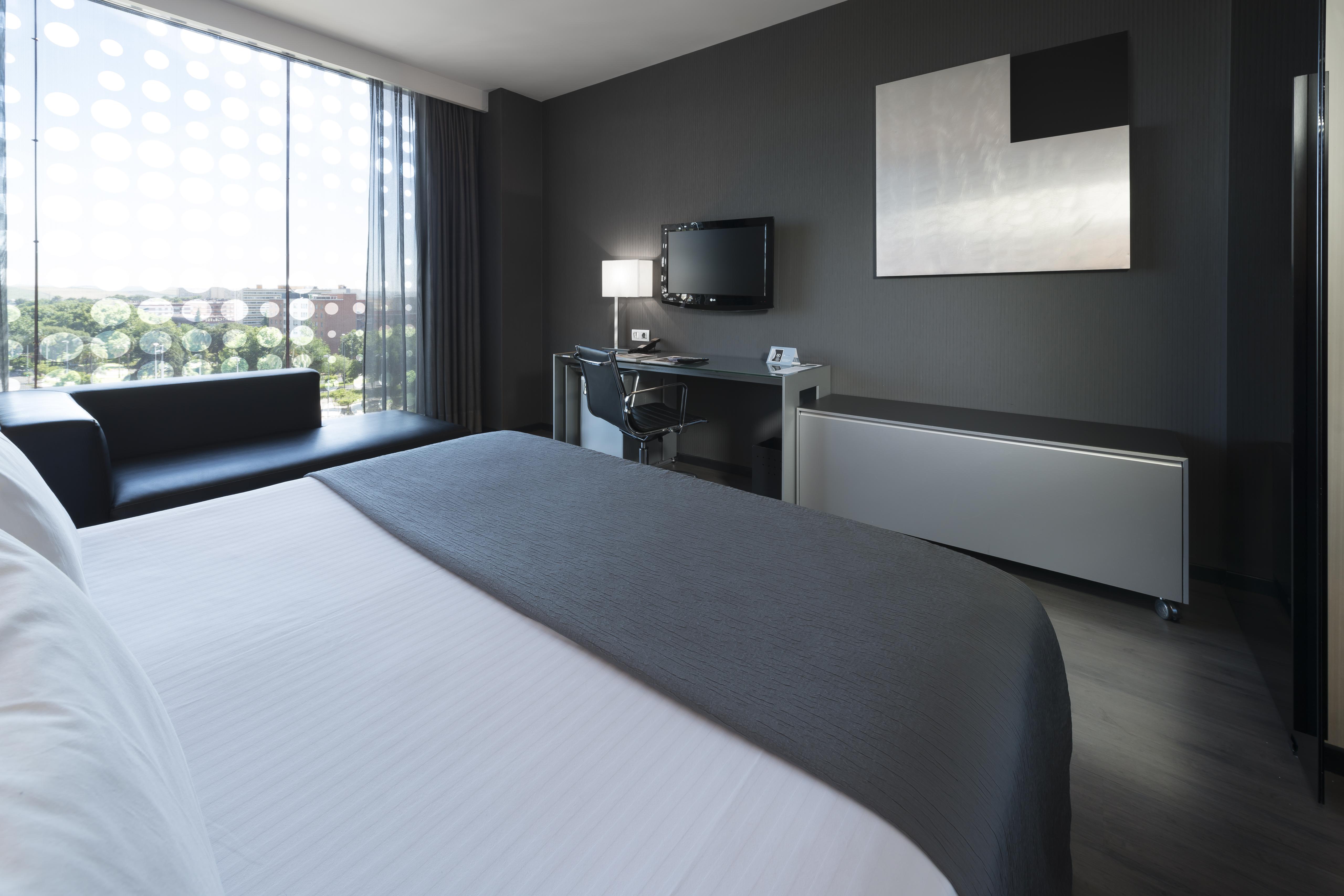 Eurostars Palace Κόρδοβα Εξωτερικό φωτογραφία The photo shows a modern hotel room featuring a sleek design. In the foreground, there is a bed with a white coverlet and gray accents. To the side, a dark sofa is visible. The room has a large window with sheer curtains, allowing natural light to en