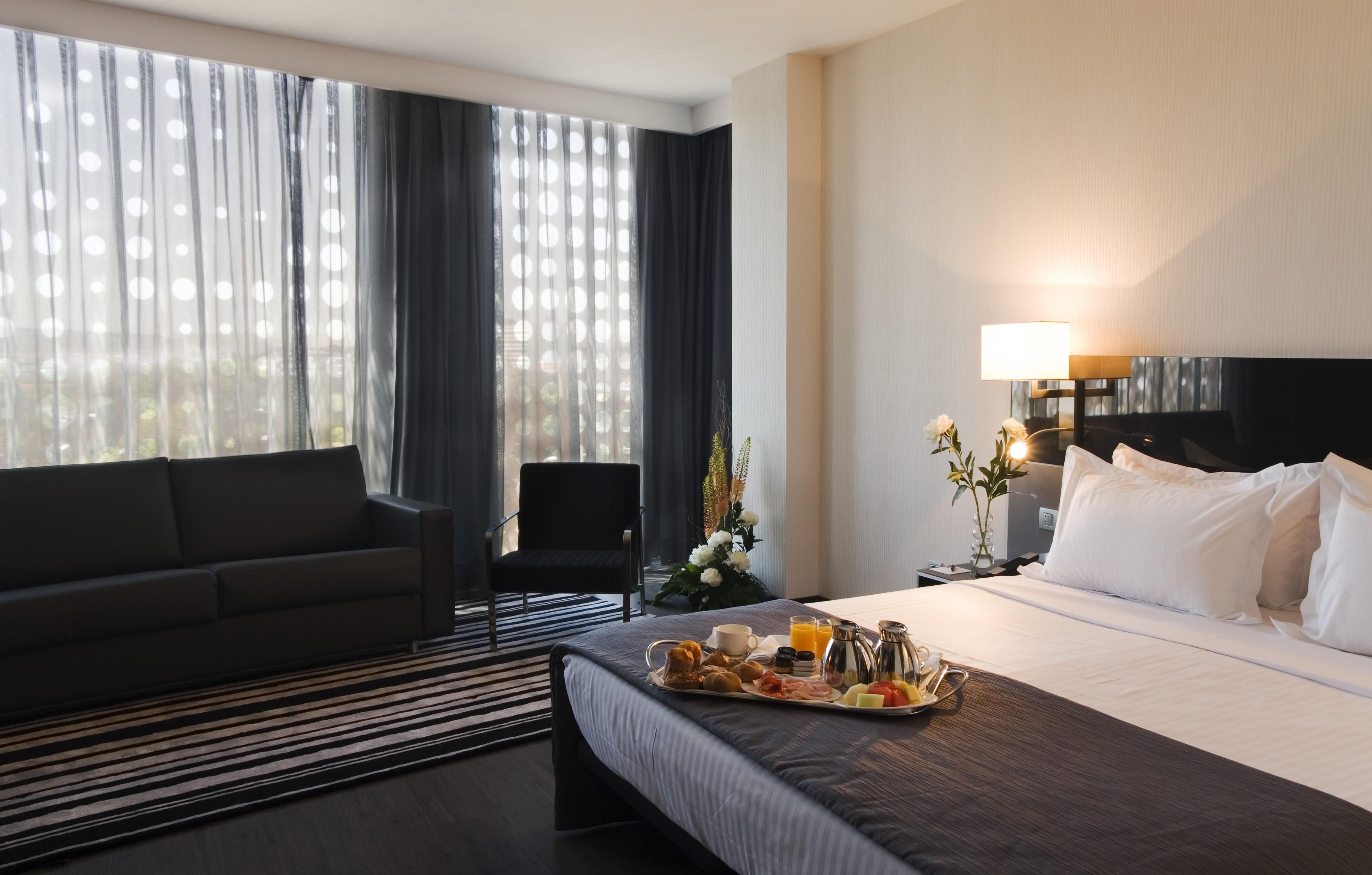 Eurostars Palace Κόρδοβα Εξωτερικό φωτογραφία The photo shows a modern hotel room featuring a neatly made bed with white linens and a dark grey blanket. There is a tray on the bed with a breakfast spread, including pastries, fruit, and beverages. To the left, there is a comfortable-looking couch