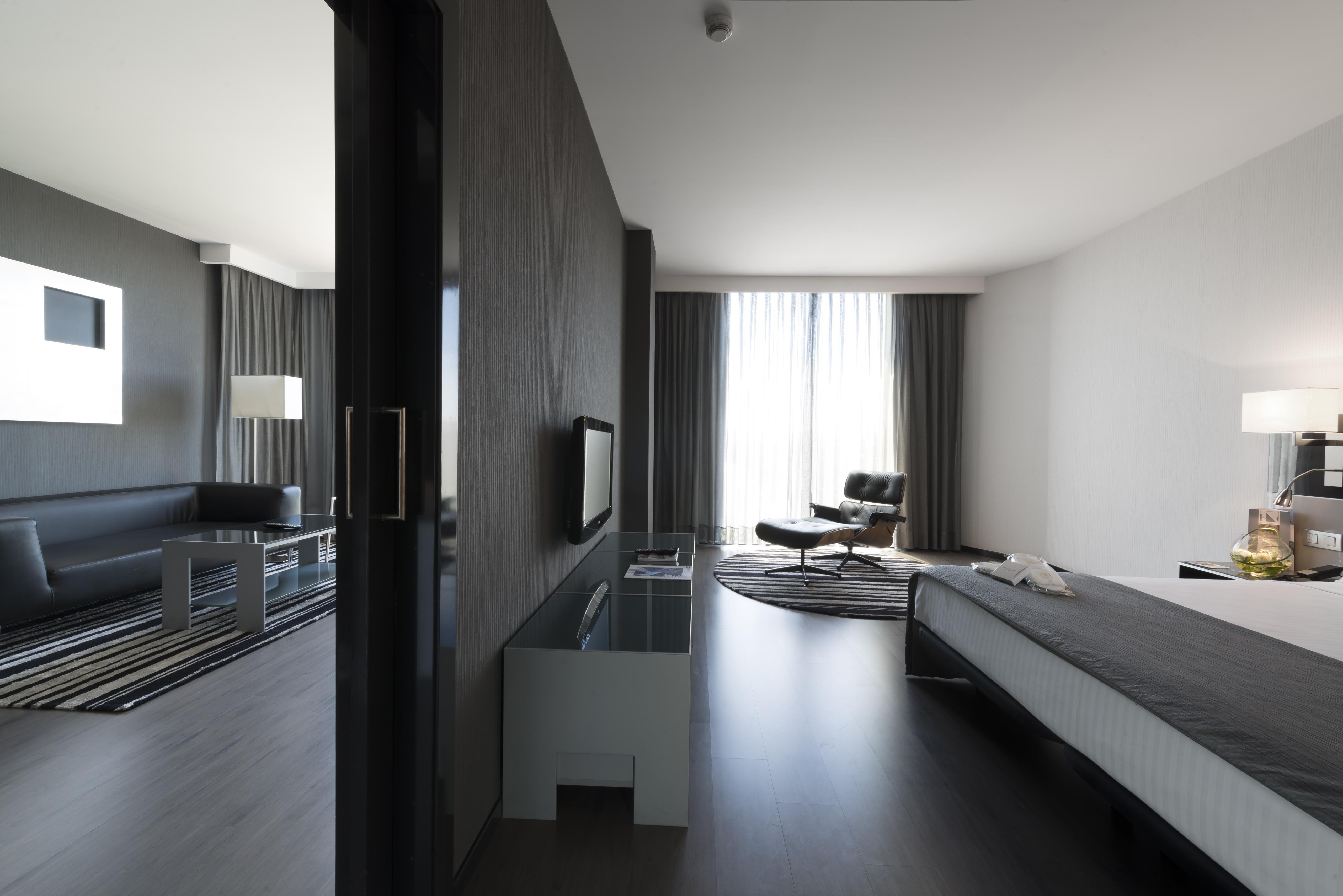 Eurostars Palace Κόρδοβα Εξωτερικό φωτογραφία The photo shows a modern hotel room or suite featuring a contemporary design. On the left side, there is a living area with a black sofa, a coffee table, and light fixtures. The large windows, covered with sheer curtains, let in natural light, enhanc