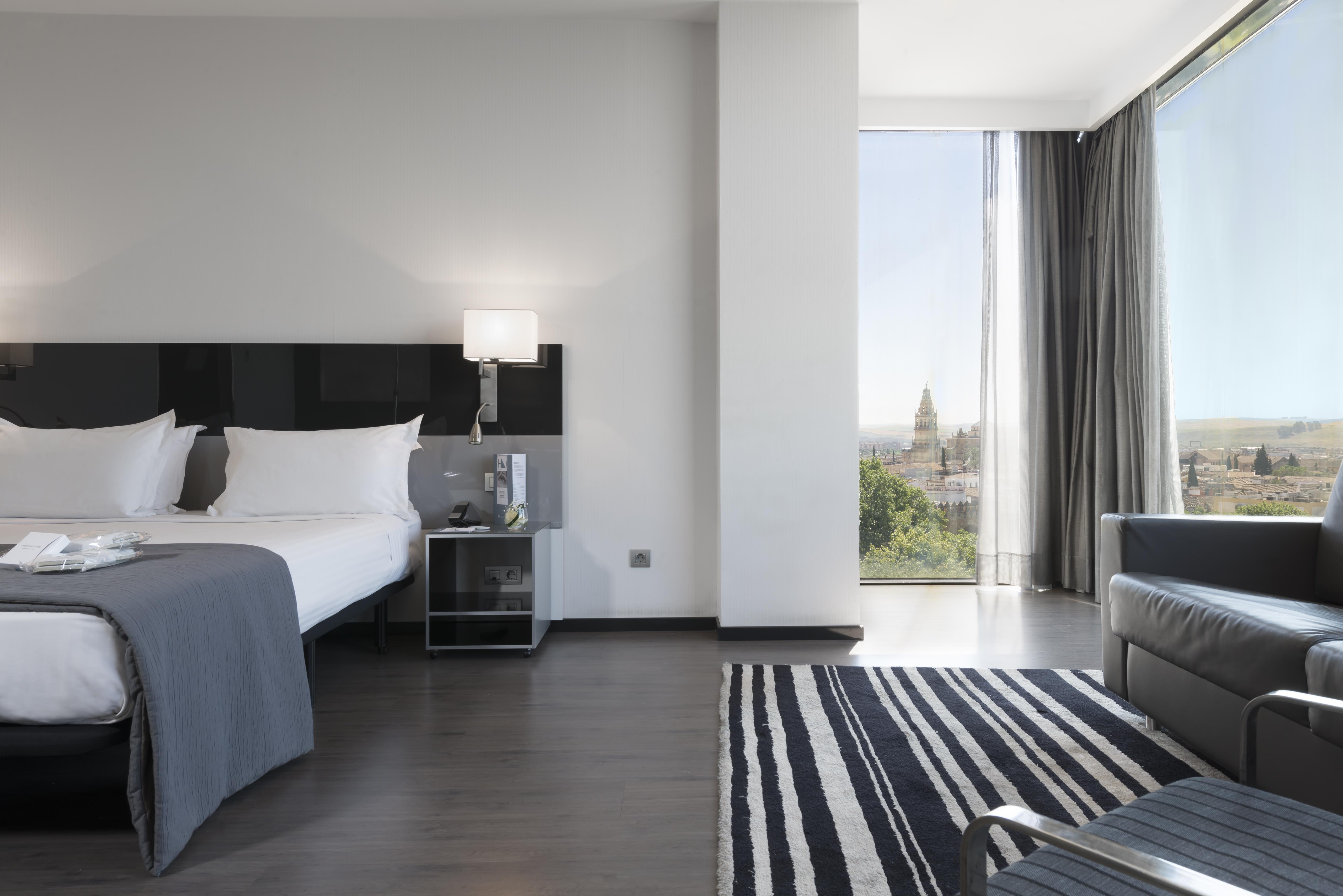 Eurostars Palace Κόρδοβα Εξωτερικό φωτογραφία The photo shows a modern hotel room featuring a large bed with white linens and dark cushions, paired with a sleek nightstand. There is a contemporary chair and a small table nearby, along with a striped area rug on the floor. A large window allows n