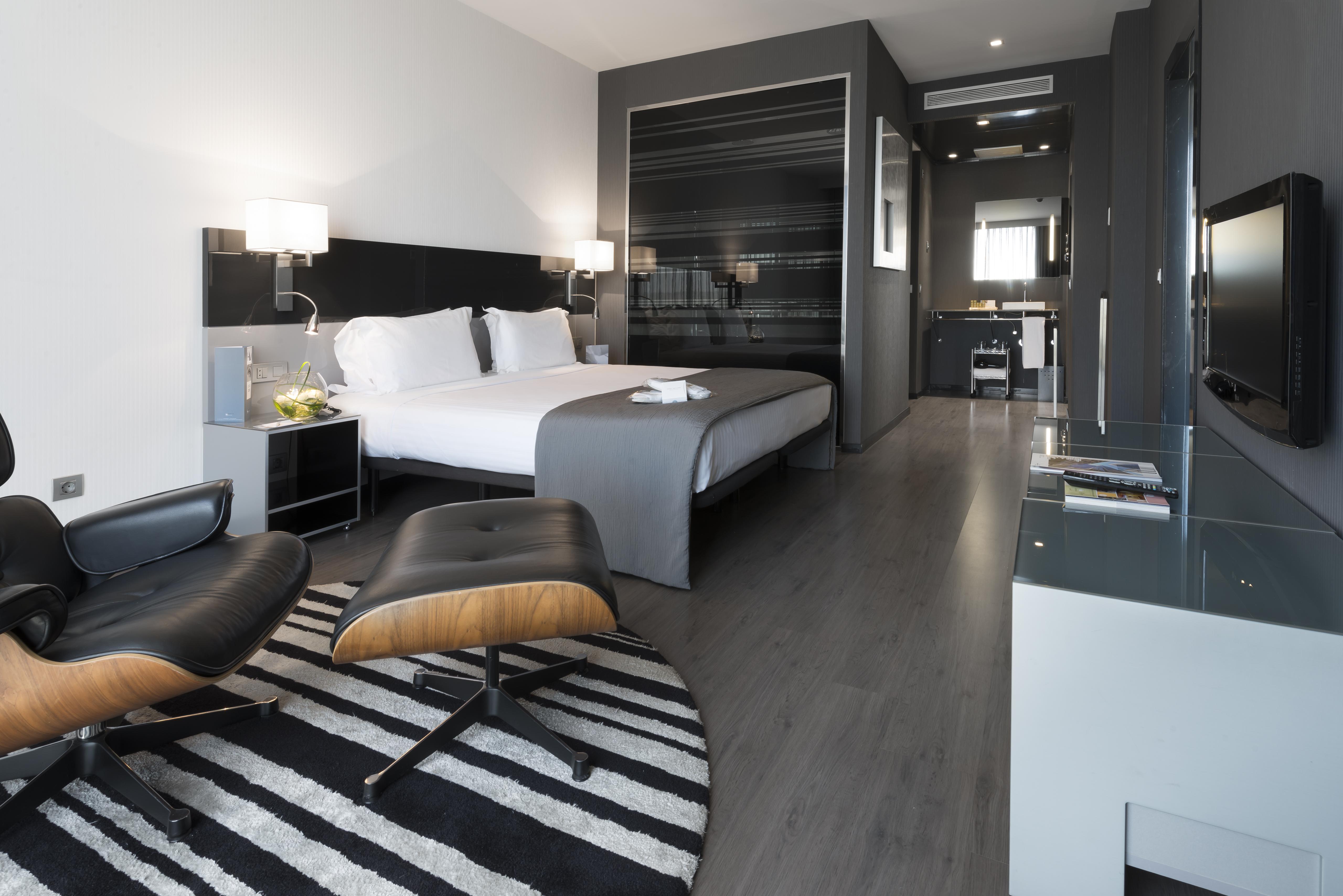 Eurostars Palace Κόρδοβα Εξωτερικό φωτογραφία The photo shows a modern hotel room featuring a king-sized bed with light-colored linens and dark accent walls. There are stylish bedside tables with lamps, and a comfortable lounge chair paired with an ottoman placed on a striped rug. The room also 