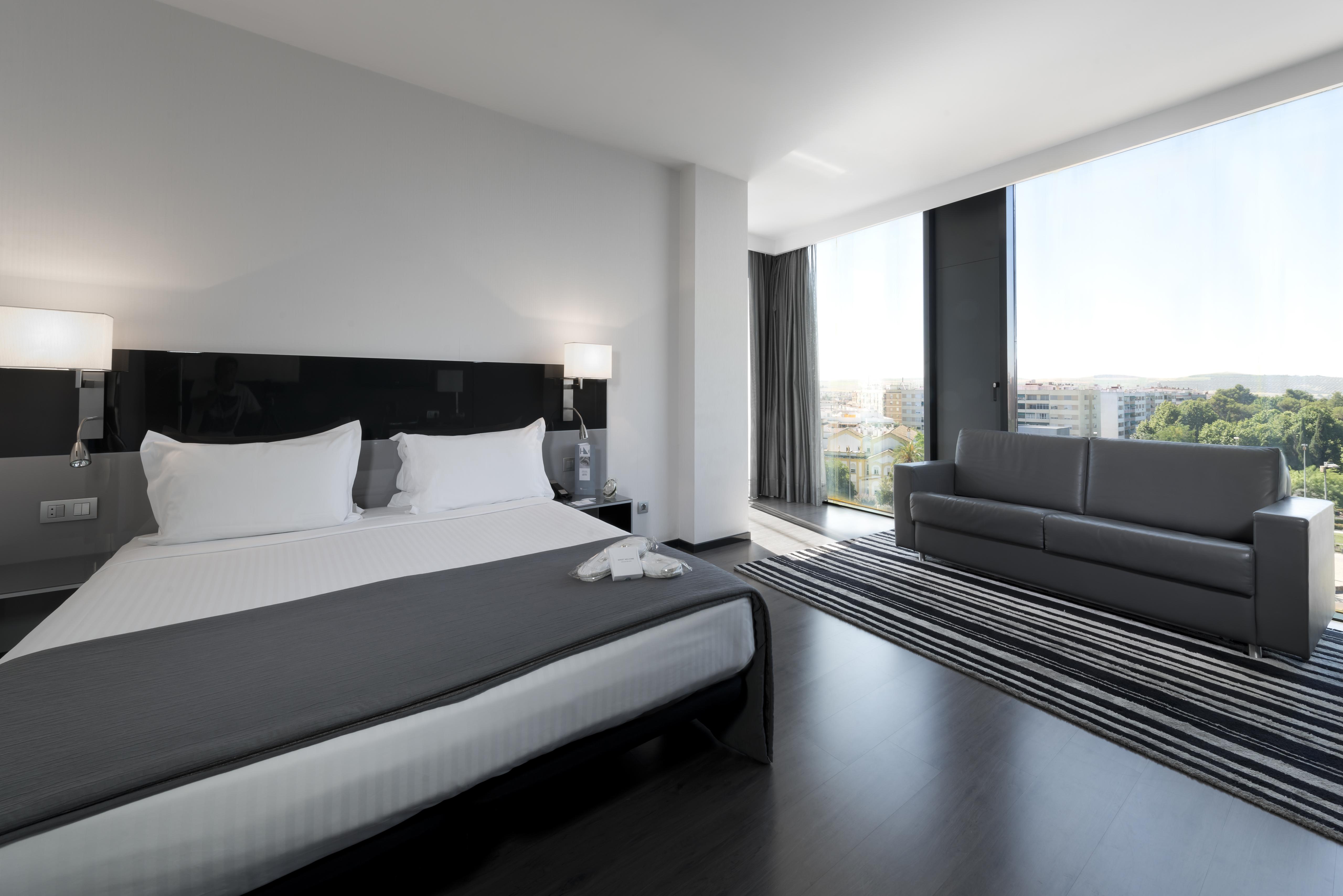 Eurostars Palace Κόρδοβα Εξωτερικό φωτογραφία The photo shows a modern hotel room with a sleek design. It features a large bed with white bedding and two pillows, accompanied by a gray throw. To the side of the bed, there are stylish nightstands with lamps. A large window allows natural light to