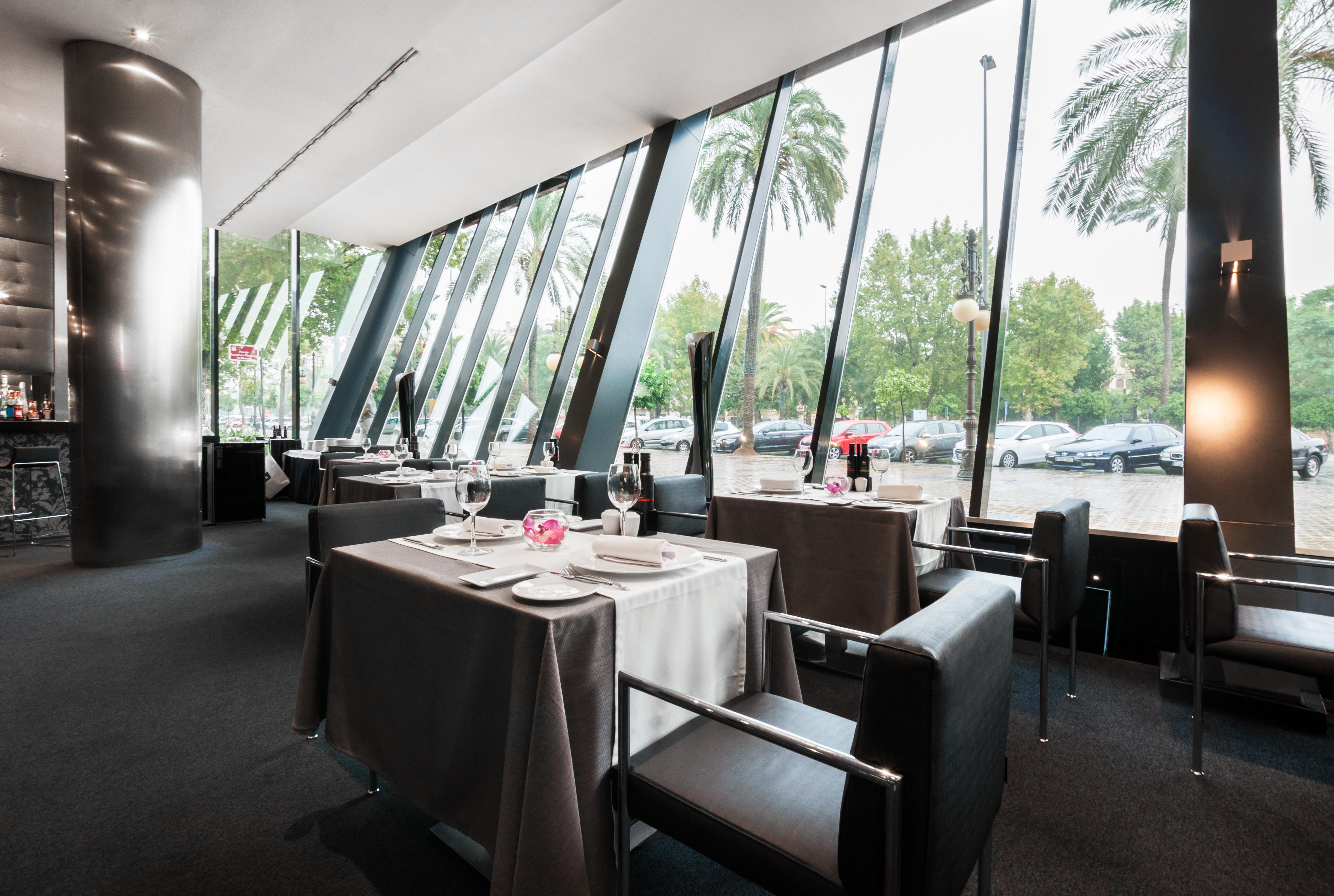Eurostars Palace Κόρδοβα Εξωτερικό φωτογραφία The photo shows the interior of a modern restaurant featuring large glass windows that provide an expansive view of the outdoors, including palm trees. The dining area is elegantly set up with tables covered in gray tablecloths, decorated with simple