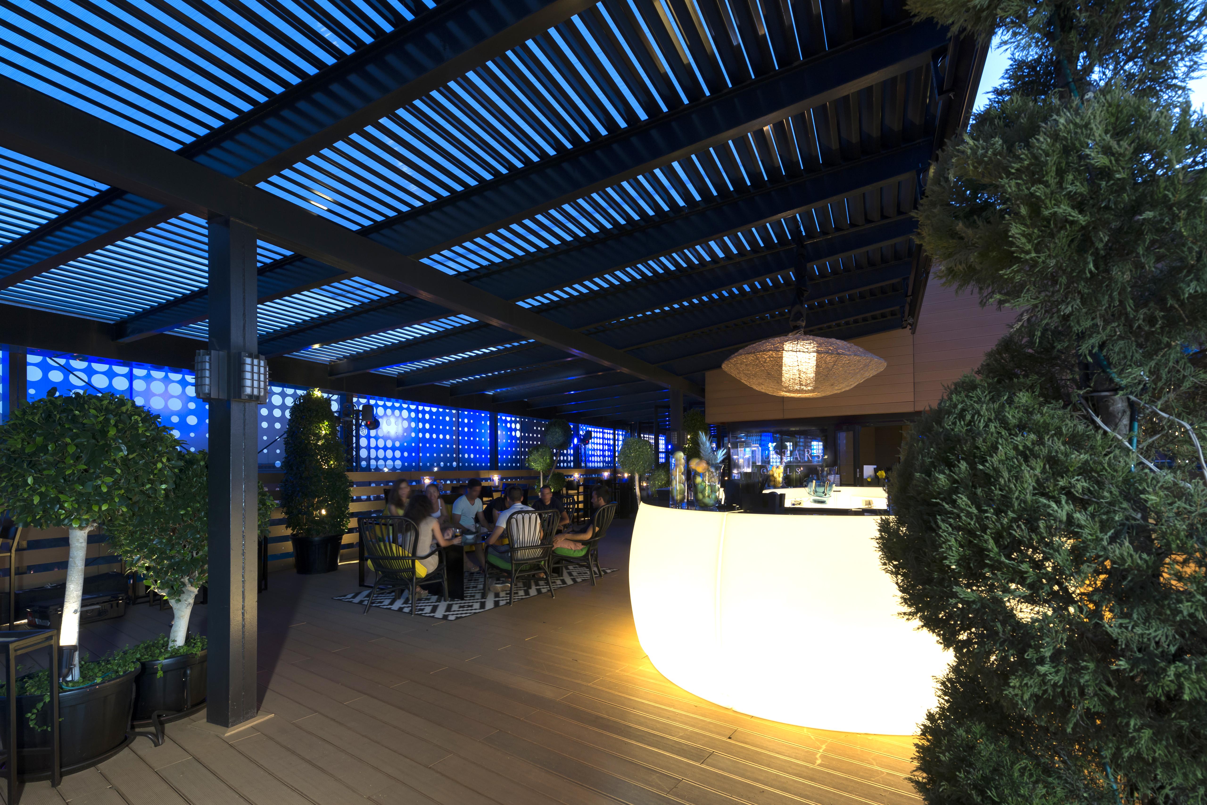 Eurostars Palace Κόρδοβα Εξωτερικό φωτογραφία The image shows a modern outdoor bar or restaurant area. The space features a sleek, illuminated bar that stands out with its curved shape. Patrons can be seen seated around tables, enjoying the ambiance. The overhead structure consists of slatted pa