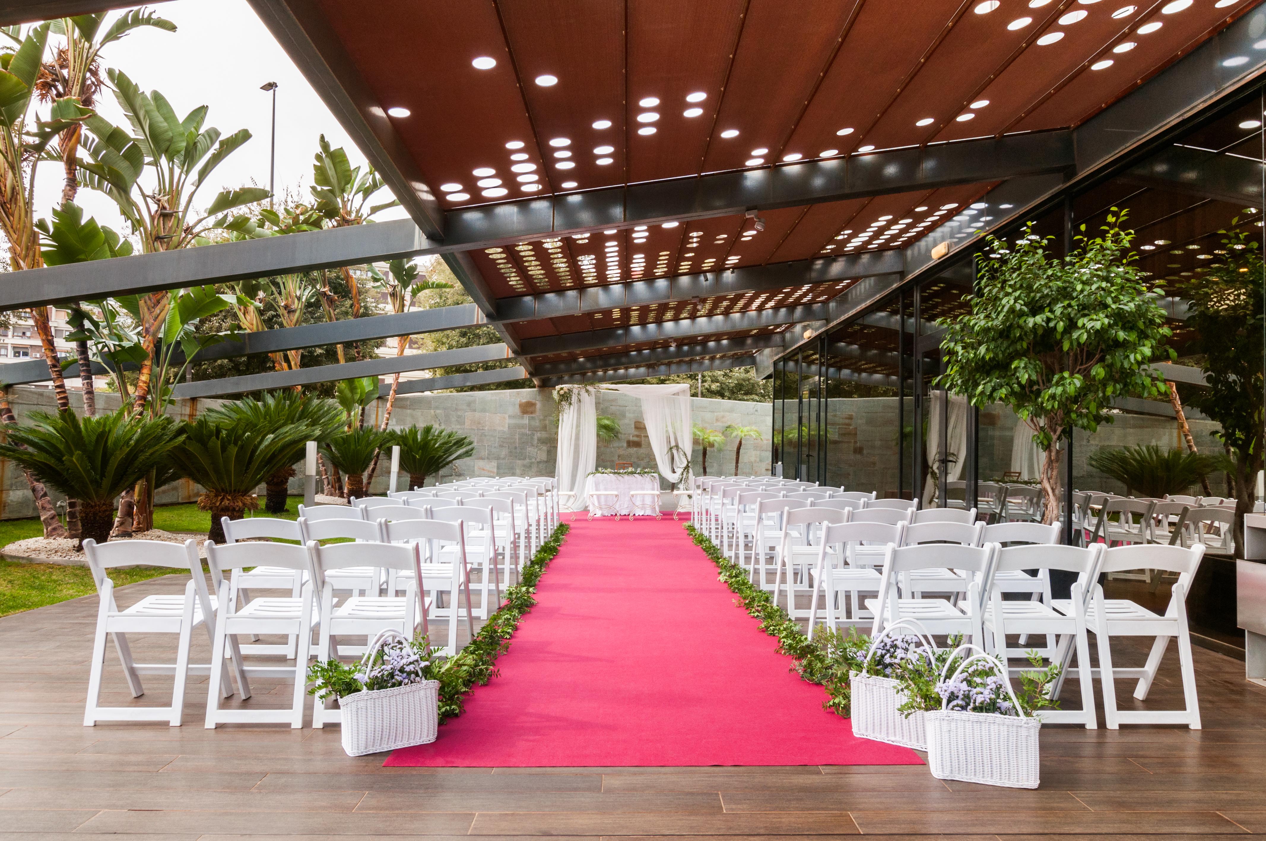 Eurostars Palace Κόρδοβα Εξωτερικό φωτογραφία The photo shows a wedding setup in an outdoor venue. There are white chairs arranged in rows on either side of a vibrant pink carpet leading to a floral arch or backdrop at the front. The area is partially shaded by a modern structure with a metal fr