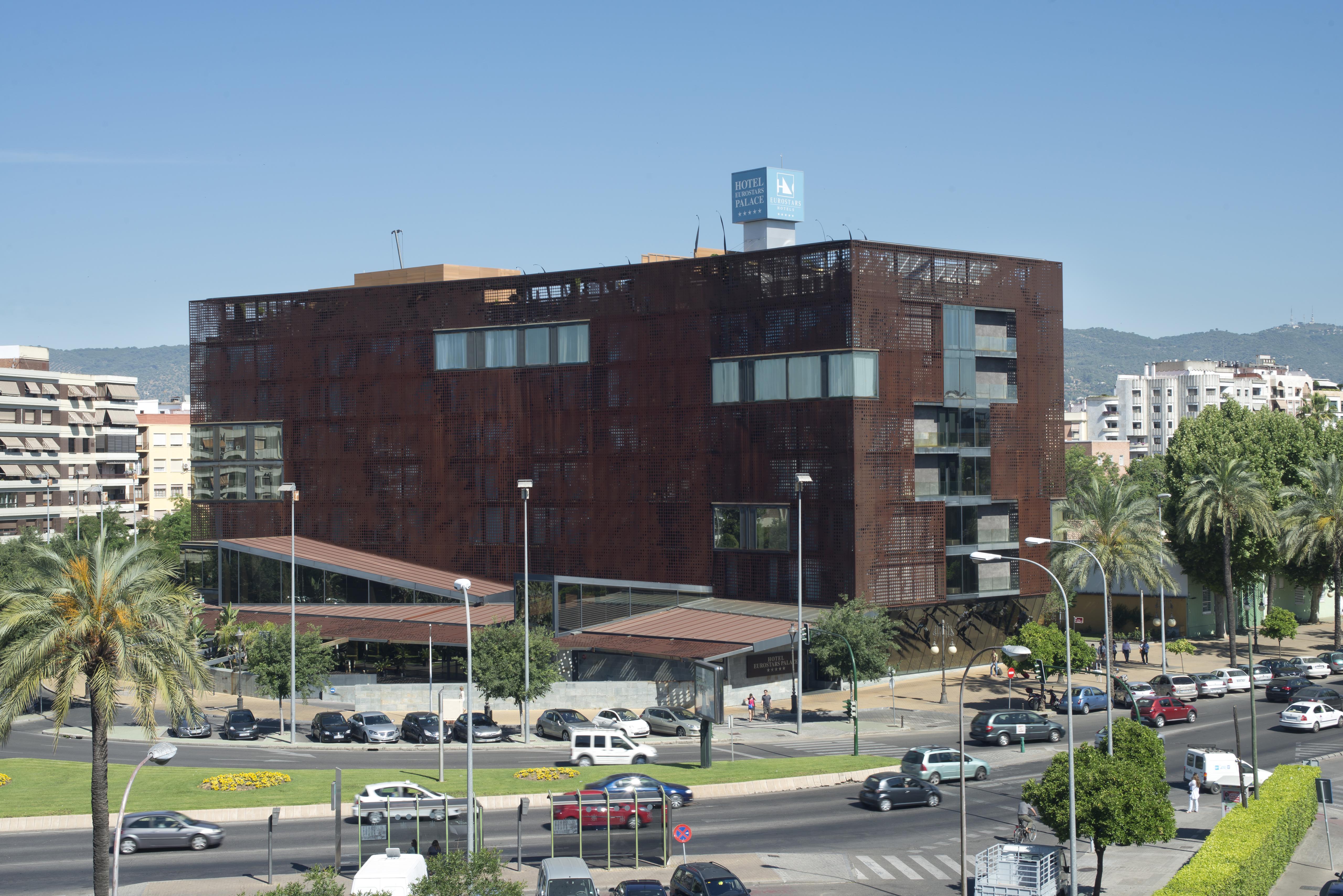 Eurostars Palace Κόρδοβα Εξωτερικό φωτογραφία The photo shows a modern building characterized by a distinctive brown, rust-like exterior, which gives it an industrial appearance. The structure has multiple large windows, allowing for natural light to enter. There are several palm trees surroundi
