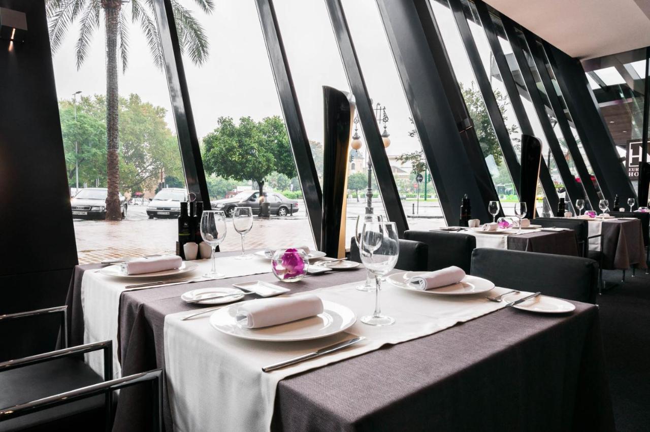 Eurostars Palace Κόρδοβα Εξωτερικό φωτογραφία The photo shows an elegant restaurant interior with large windows featuring modern angular designs. There are two dining tables set up, each covered with white and dark tablecloths. The tables are elegantly arranged with plates, cutlery, and wine gla
