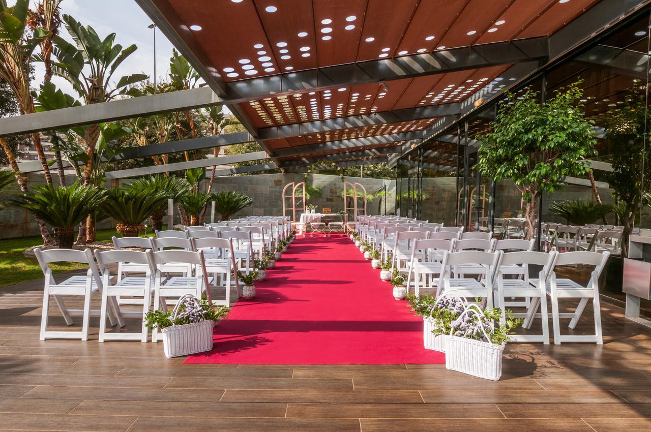 Eurostars Palace Κόρδοβα Εξωτερικό φωτογραφία The image shows an outdoor wedding setup. There are white chairs arranged neatly on either side of a vibrant red carpet that leads to a framed archway. The area is covered by a glass and metal structure, allowing plenty of natural light, while tropic