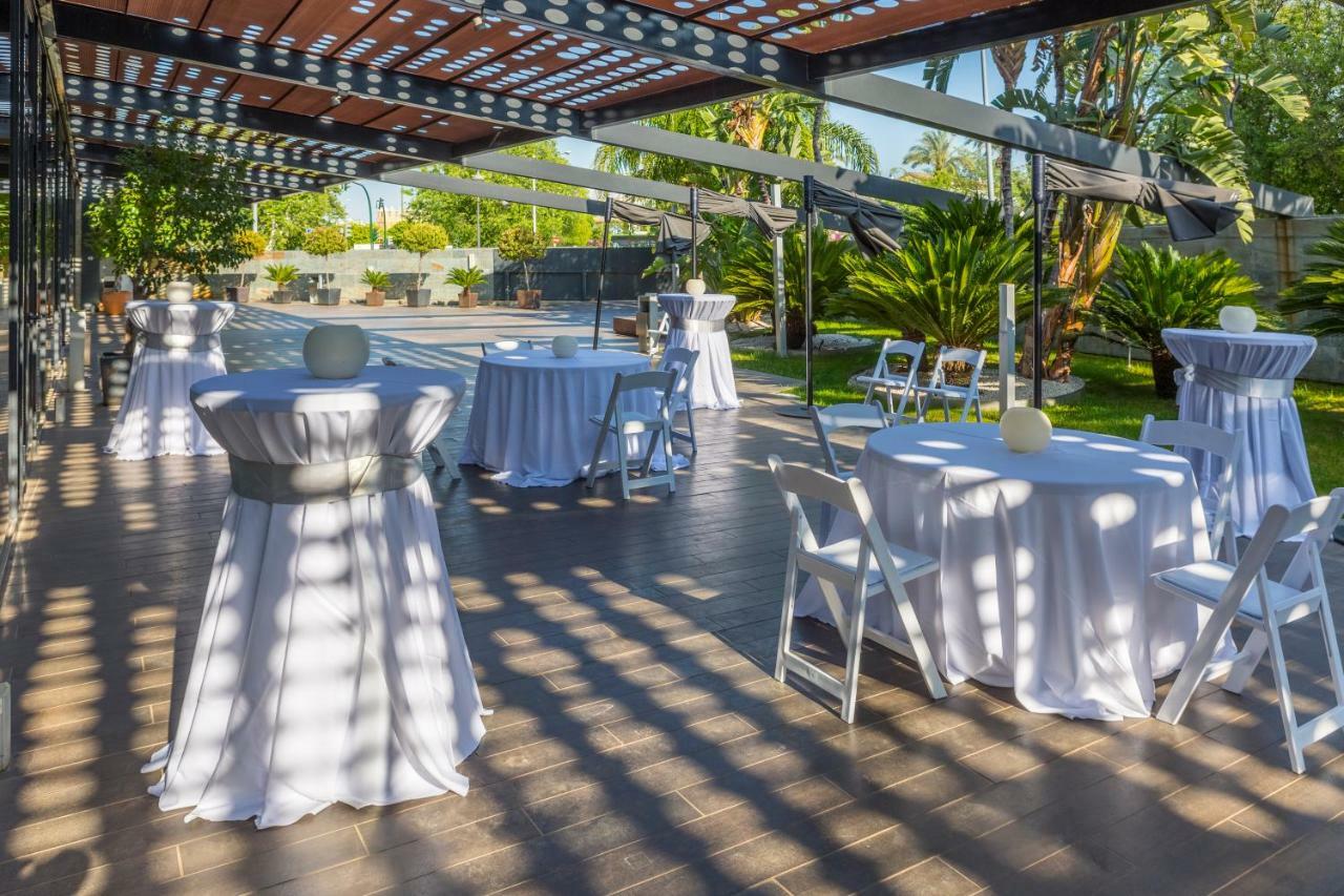 Eurostars Palace Κόρδοβα Εξωτερικό φωτογραφία The photo shows an outdoor event setup. There are several round tables covered with white tablecloths, some adorned with decorative elements like small centerpieces. Chairs are arranged around the tables, and the area is surrounded by greenery, inclu